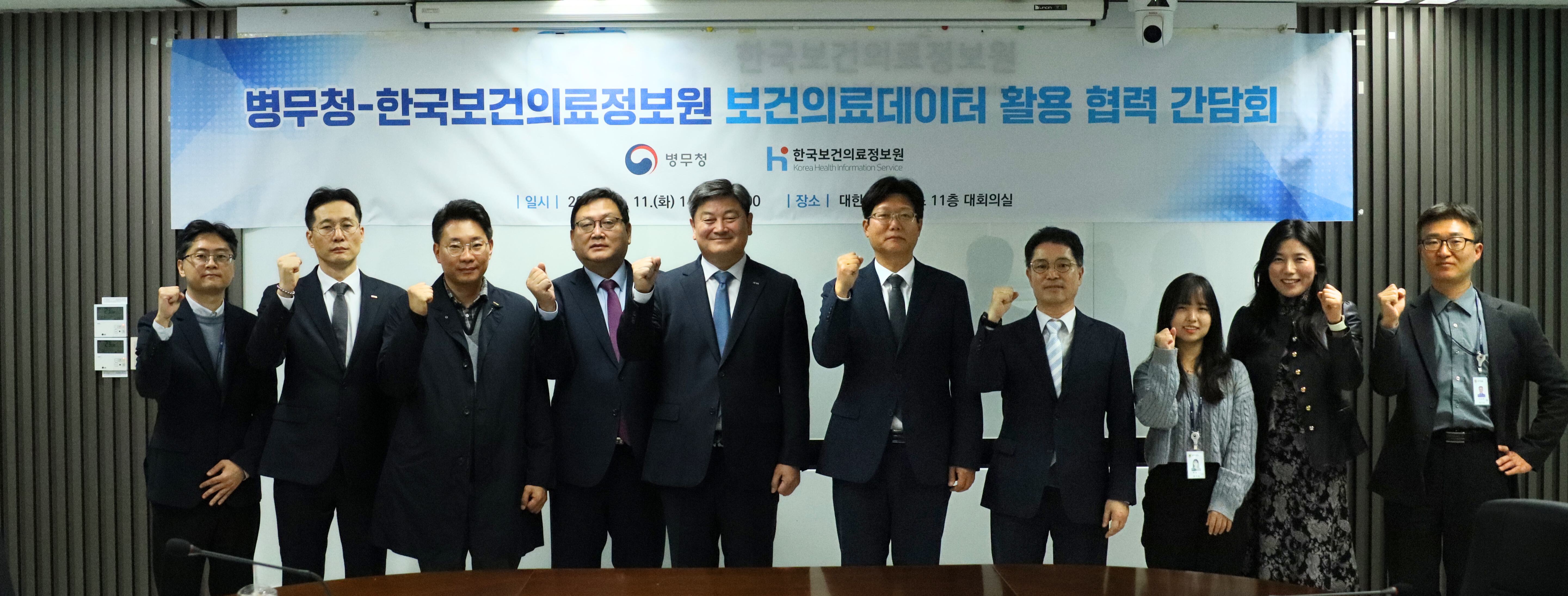 사진에는 회의실 전면에 설치된 현수막을 배경으로
양 기관 관계자들이 일렬로 서서 주먹을 쥔 포즈로 기념 촬영을 하고 있다.

배경 현수막에는
‘병무청-한국보건의료정보원 보건의료데이터 활용 협력 간담회’라는 문구와 함께
병무청 및 한국보건의료정보원(KHIS) 로고가 함께 표기되어 있다.
현수막 하단에는 간담회 개최 일시와 장소 정보가 기재되어 있어
공식적인 협력 행사임을 알 수 있다.
