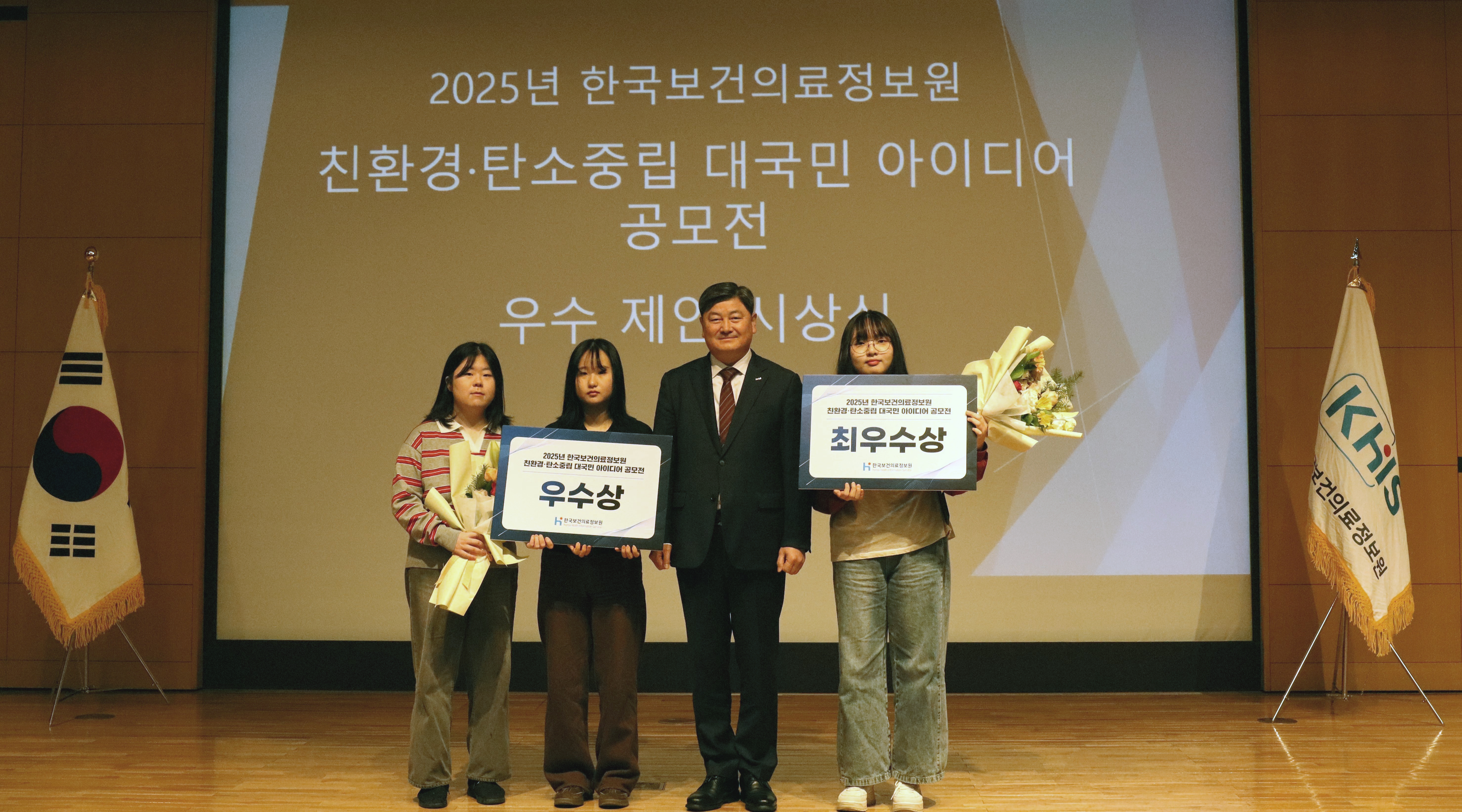 친환경·탄소중립 대국민 아이디어 공모전 시상식 개최 사진 2025년 한국보건의료정보원 친환경·탄소중립 대국민 아이디어 공모전 우수 제안 시상식에서 수상자들이 무대 위에 서서 상장과 꽃다발을 들고 기념촬영을 하는 모습. 가운데에는 한국보건의료정보원장이 정장을 입고 서 있으며, 양옆에는 ‘우수상’과 ‘최우수상’ 수상자가 상패를 들고 있다. 무대 뒤 대형 화면에는 공모전 명칭과 시상식 안내 문구가 표시되어 있고, 좌우에는 태극기와 한국보건의료정보원 깃발이 배치되어 있다.