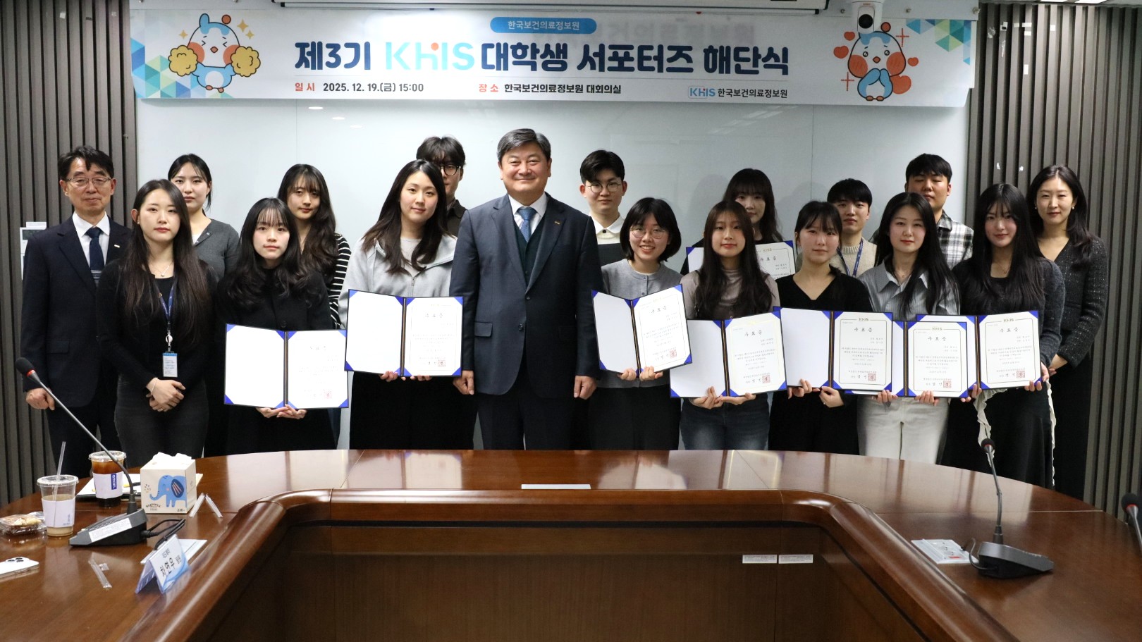 한국보건의료정보원 회의실에서 열린 제3기 대학생 서포터즈 해단식 현장에서 단체 기념촬영을 하고 있다. 중앙에는 한국보건의료정보원 관계자가 정장을 입고 서 있으며, 양옆으로 대학생 서포터즈들이 줄지어 서서 수료증을 펼쳐 들고 있다. 뒤편에는 ‘제3기 KHIS 대학생 서포터즈 해단식’이라는 현수막이 걸려 있고, 회의용 테이블과 마이크가 보이는 공식 행사 공간에서 대학생들의 홍보 활동 성과와 참여를 마무리하는 자리임을 보여준다.