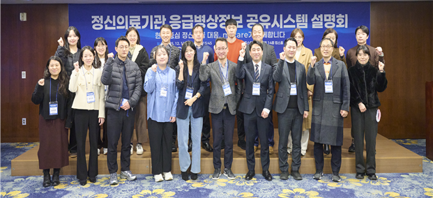 ‘정신의료기관 응급병상정보 공유시스템 설명회’ 현장에서 관계자들이 단체 기념촬영을 하고 있다. 행사장 전면에는 ‘정신의료기관 응급병상정보 공유시스템 설명회’라는 문구가 적힌 현수막이 걸려 있으며, 참석자들은 정장 또는 캐주얼 복장에 명찰을 착용한 채 주먹을 쥐고 결의를 다지는 포즈를 취하고 있다. 이 사진은 정신응급 대응 강화를 위해 응급병상정보 공유시스템(m-care)의 전국 운영을 알리는 공식 행사 분위기와 협력 의지를 보여준다.